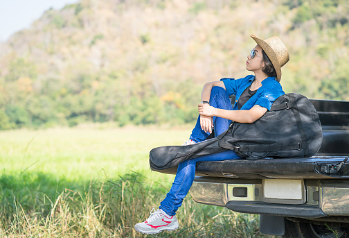 Young asian women short hair wear hat and sunglasses carry her guitar bag ,sit on pickup truck in countryside Thailand