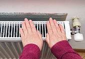 Senior woman trying to keep warm by warming hands on the heating radiator in winter time