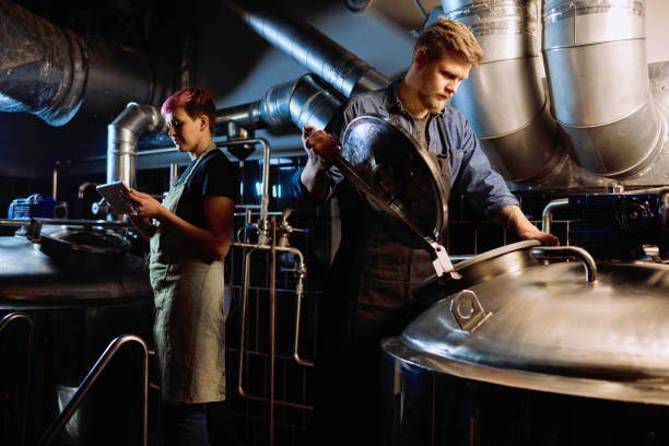 junge blonde bärtigen männlichen brauer in arbeitskleidung öffnung deckel von riesigen stahltank - brauerei stock-fotos und bilder