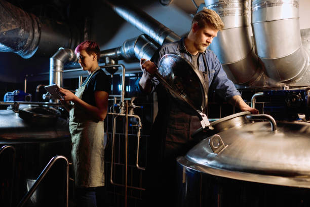 Young blond bearded male brewer in workwear opening lid of huge steel tank Young blond bearded male brewer in workwear opening lid of huge steel tank for beer preparation while working with female colleague distillery photos stock pictures, royalty-free photos & images