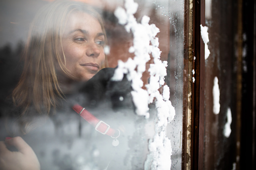 A pretty woman is looking through a snowy window and smirking while hugging her dog