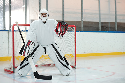 Hockey player in white sports uniform, protective helmet and gloves holding stick while standing on rink against net and waiting for puck