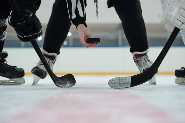 mano dell'arbitro che tiene il disco sulla pista di pattinaggio con due giocatori in piedi intorno a lui - hockey su ghiaccio foto e immagini stock