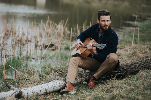 caucasian bearded man is playing guitar near the lake. handsome stylish hipster.