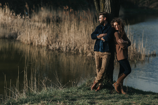 two caucasian lovers are standing near tree by the lake. Young couple is hugging on autumn day outdoors. A bearded man and curly woman in love. Valentine's Day. Concept of love and family