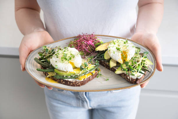 Girl holding a plate with toast made with microgreens, poached eggs, avocado, asparagus and cheese. Closeup background. Girl holding a plate with toast made with microgreens, poached eggs, avocado, asparagus and cheese. Closeup background. eating asparagus stock pictures, royalty-free photos & images