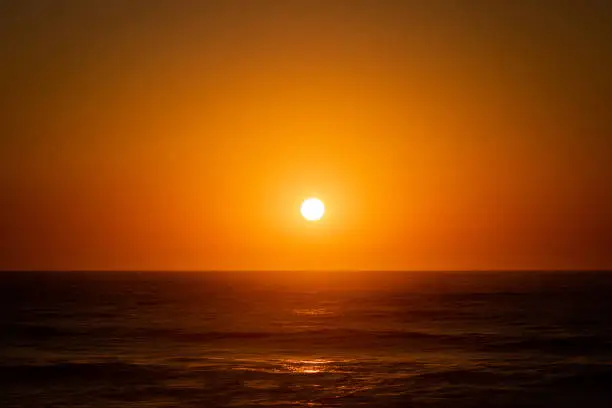 The bright round sun setting above the calm Atlantic Ocean and illuminating the whole clear sky and water in Quiaios Beach, Portugal. Nature background with the sunset and sea