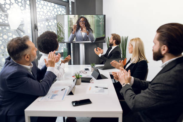 istock Videoconference, telemeeting, remote work. Multiethnic diverse business team, sitting around the table in office room, greeting their female African colleague, clapping hands and looking at tv screen 1297306625