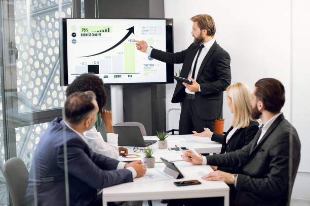 istock Front view of young confident bearded businessman, showing statistic datas on big plasma screen for his focused high-skilled multiethnical colleagues during meeting in office room 1297307672