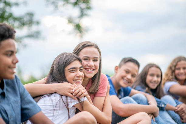 A multi ethnic group of teenage boys and girls hanging outdoors at a park. They are all sitting down on grass.