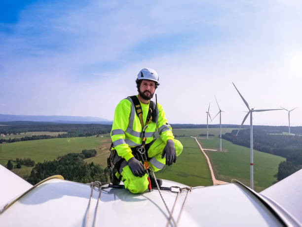 Industrial climber,  rope access technician kneeling on enormous onshore wind-turbine while preparing for blade inspection with wind farm behind him stock photo