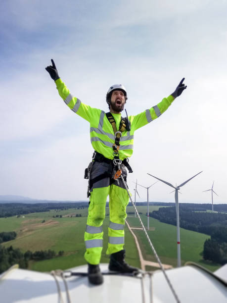 istock Handsome Industrial climber,  rope access technician standing on enormous onshore wind-turbine, hand raised and screaming - celebrating before abseil on blade with wind farm behind and epic view 1297308943