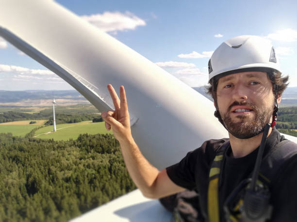 Happy Industrial climber, rope access technician is smiling and took amazing selfie with helmet and harness on gigantic onshore wind-turbine hub and blade and view behind him and hand victory gesture feliz escalador industrial, técnico de acceso a la cuerda está sonriendo y tomó un selfie increíble con casco y arnés en el gigantesco cubo de turbina de viento en tierra y la hoja y la vista detrás de él y gesto de la victoria de la mano