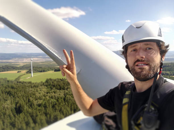 Happy Industrial climber,  rope access technician is smiling and took amazing selfie with helmet and harness on gigantic onshore wind-turbine hub and blade and view behind him and hand victory gesture Happy Industrial climber,  rope access technician is smiling and took amazing selfie with helmet and harness on gigantic onshore wind-turbine hub and blade and view behind him and hand victory gesture czech republic photos stock pictures, royalty-free photos & images