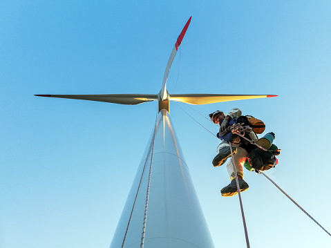 Low angle view on Rope access technician, industrial climber hanging in the rope on wind turbine enlightened by the sunset and looking looking into the camera with shoe sleeves