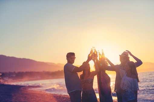 Group of young people partying on the beach at sunset. They are all holding bottles of beer and celebrating with a toast. Silhouette with the beach, sun and sea in the background.