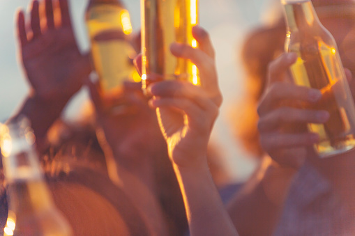 Group of young people partying on the beach at sunset. They are all holding bottles of beer and celebrating. Tight crop with close up of hands.