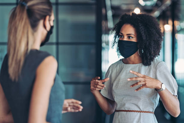Businesswomen talking in the office during the covid-19 pandemic Businesswoman, wearing a mask, standing at distance and talking to a colleague new normal concept stock pictures, royalty-free photos & images