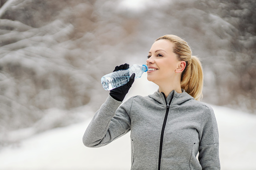 Sportswoman drinking water and taking a break while standing in nature at snowy winter day. Healthy lifestyle, winter fitness