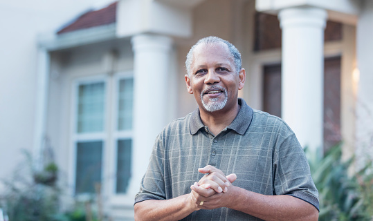 A senior African-American man standing in front of his house. He is a retired homeowner, smiling at the camera. He is in his 60s.