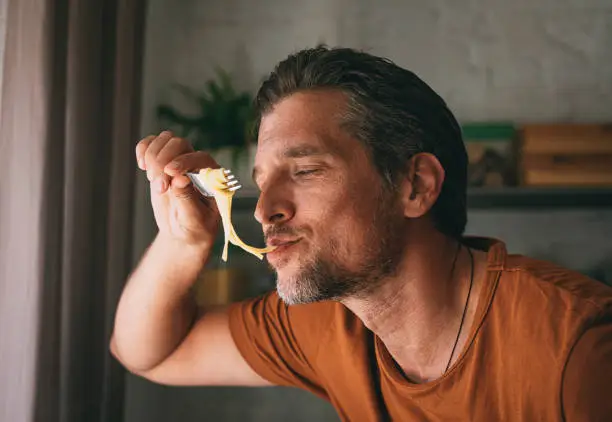 Close up shot of a handsome young happy Caucasian man tasting pasta with a fork while preparing a meal in a kitchen.