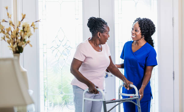 Home nurse helping senior woman at home using walker A healthcare worker visiting a senior African-American woman at home, helping her use a mobility walker. The patient is in her 70s and the nurse is a mature woman in her 40s. home caregiver stock pictures, royalty-free photos & images