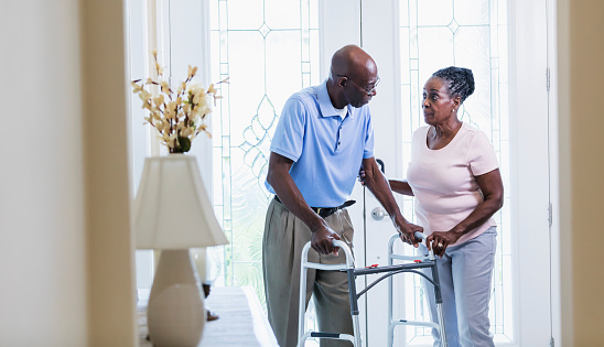 A senior African-American couple inside their home, at the front door. The man is using a mobility walker and his wife is helping him. They are in their 70s.