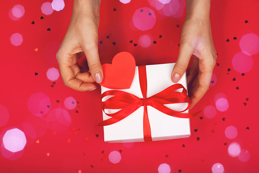 Beautiful female hand with minimalistic gift on a red background with cute heart. Concept of the Valentine's Day, surprises with love, holiday, etc.