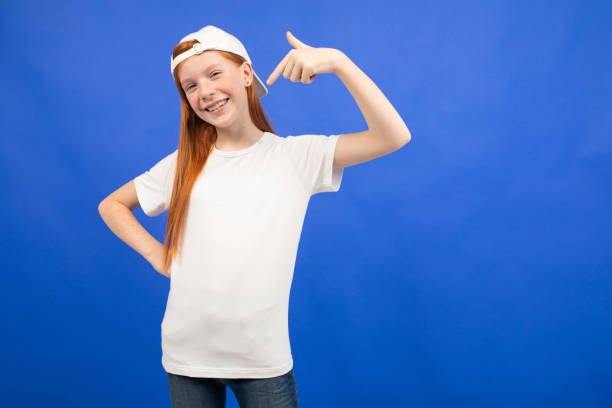 attraente ragazza adolescente dai capelli rossi in una t-shirt bianca con un layout mostra la classe su uno sfondo blu studio - top of the class foto e immagini stock
