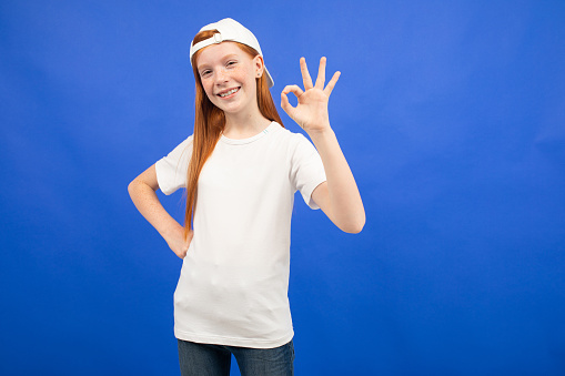 charming red-haired teenager girl in a white T-shirt shows a blank print space on a blue studio background.