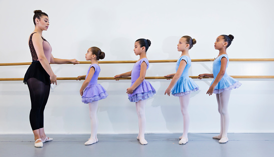 A young Hispanic woman in her 20s giving ballet lessons to a multi-ethnic group of four girls, 8 to 10 years old. The students are standing in a row, facing the teacher, doing barre work.