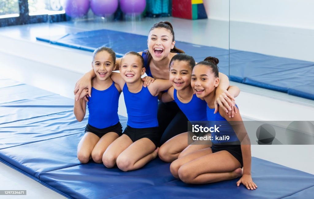 Gymnastics coach with four girls in gym A young Hispanic woman coaching a multi-ethnic group of four gymnasts. The girls are 8 to 10 years old. They are sitting together on a mat in the gym, smiling at the camera, and then looking at their coach. Gymnastics Stock Photo