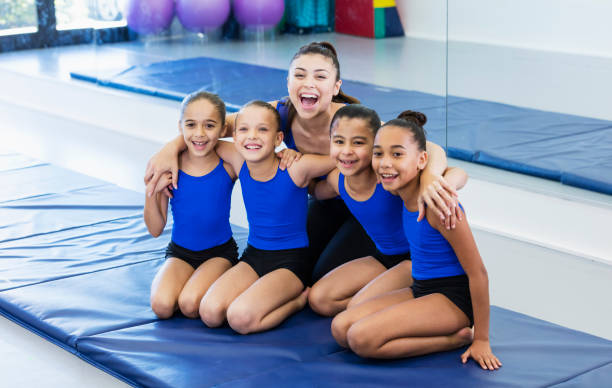 Gymnastics coach with four girls in gym A young Hispanic woman coaching a multi-ethnic group of four gymnasts. The girls are 8 to 10 years old. They are sitting together on a mat in the gym, smiling at the camera, and then looking at their coach. gymnastics stock pictures, royalty-free photos & images