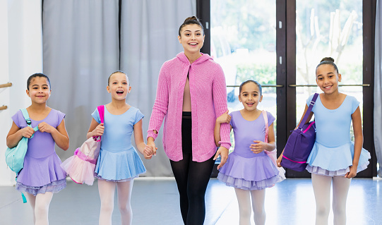 A multi-ethnic group of four ballet students and their teacher arriving at a dance studio together, holding hands, carrying tote bags. The instructor is a young Hispanic woman in her 20s. The girls are 8 to 10 years old. They are smiling at the camera.