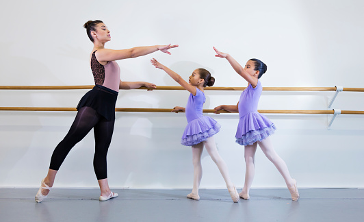 A young Hispanic woman in her 20s giving ballet lessons to two Hispanic 8 year old girls, doing barre work. The students are facing the teacher, holding the barre, reaching up gracefully with one arm, fingers and toes pointed.
