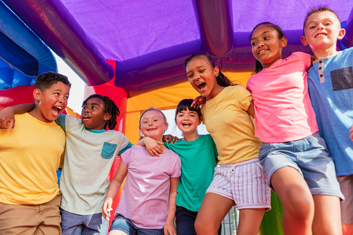 A multi-ethnic group of seven children standing together in a bounce house, side by side, smiling and looking at the camera. They are 8 to 10 years old. The girl third from left next to the African-American boy, has down syndrome.