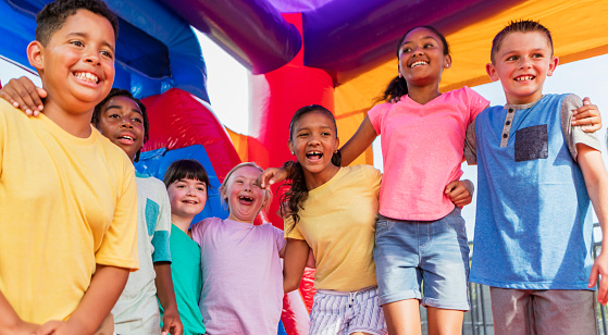 A multi-ethnic group of seven children standing together in a bounce house, side by side, smiling. They are 8 to 10 years old. The girl in the middle has down syndrome.
