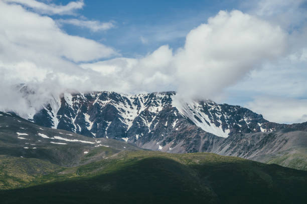 istock Awesome mountains landscape with big snowy mountain ridge among low clouds in blue sky. Atmospheric highland scenery with high mountain range with snow above sunlit green hill. Beautiful view to rocks 1297359920