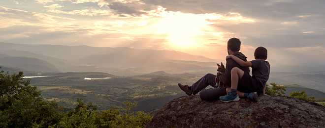 Father, son and dog on top of rock against sunset over Rhodope mountains, Bulgaria. Family spends happy time together in nature. Local tourism, social distancing, hiking. Panorama, banner, copy space