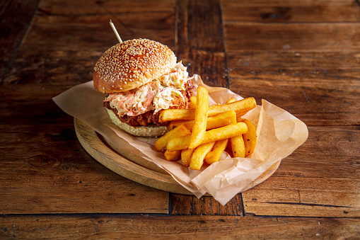 Brioche bun with sesame on top, breaded chicken burger garnished with coleslaw salad and served with french fries on wooden plate