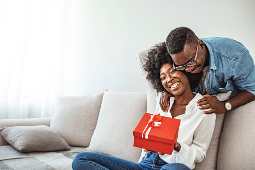 Xmas surprise. Afro man giving Christmas present to girlfriend. Thankful afro woman with christmas gift hugging husband at home. Presenting gift as Christmas eve tradition