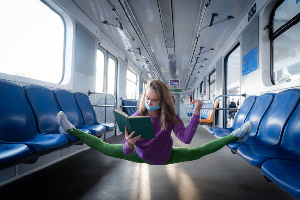 非常に柔軟なマスクの女性は、体操のスプリットに座って地下鉄の車の中で本を読みます。健康的なライフスタイル、柔軟性、ヨガのコンセプト - leg split ストックフォトと画像
