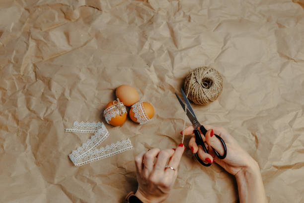 istock Decorating easter eggs on kraft paper with lace and rope. Three easter eggs on kraft paper background. Woman cuts the rope with black scissors. 1297371807