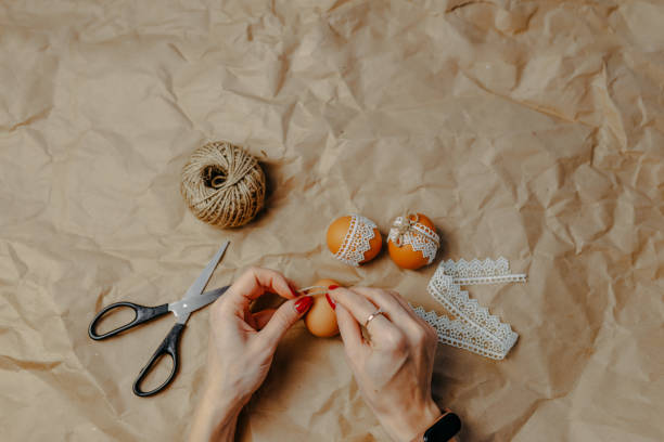 istock Decorating easter eggs on kraft paper with lace and rope. Three easter eggs on kraft paper background. Woman binding rope around the egg. 1297371829