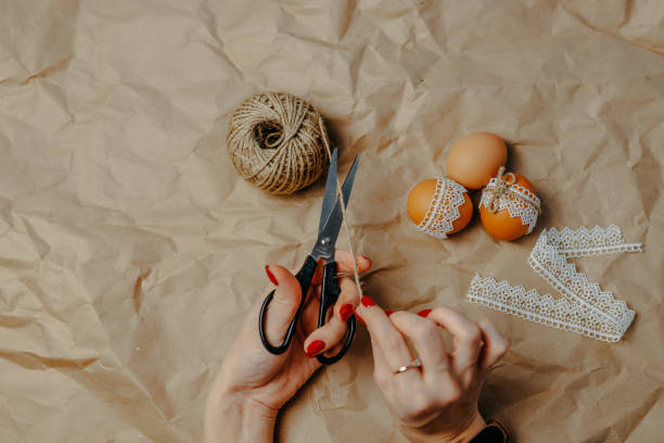 istock Decorating easter eggs on kraft paper with lace and rope. Three easter eggs on kraft paper background. Woman cuts the rope with black scissors. 1297371830