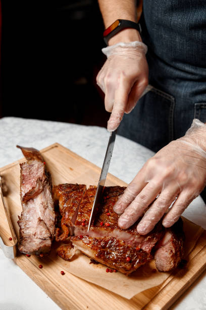 istock Waiter with gloves cuts Tomahawk steak on a beef bone into pieces for restaurant guests. Close-up of hands 1297372489