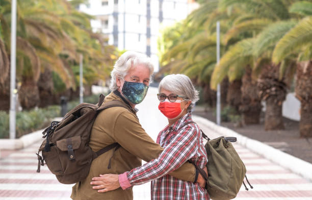 istock Back view of two white-haired traveler people looking at camera while walking together during the city tour, wearing a surgical mask due to the coronavirus Active retired people enjoying travel and freedom 1297375268