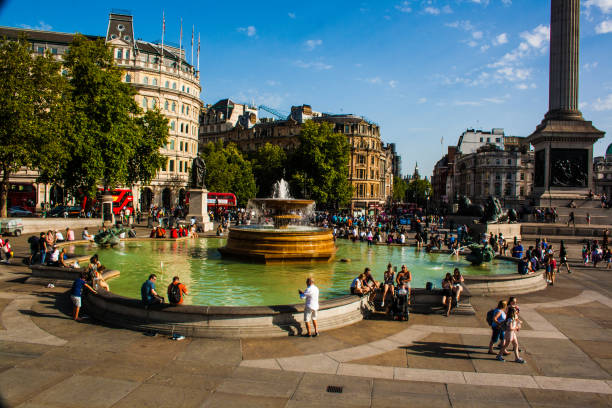 A senior adult black female tourist leaning on a fountain in London. She is located on the big Trafalgar square and looking around admiring the city. She is taking a break after a long day of visiting landmarks in London. The weather is cold and gloomy.