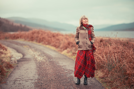 Mature woman wearing a red tartan kilt and scarf on a cloudy winter's day at Loch Arkaig in the Highlands of Scotland.