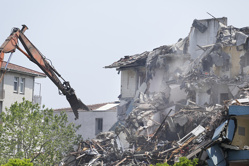 Behind the dilapidated brick wall of the building, excavators are demolishing the house against the background of a bright sky